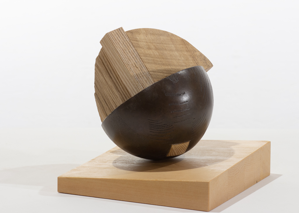 Sculpture composite faite de bronze et de bois. De forme ronde, elle est posée sur un plan incliné afin de la faire basculer légèrement.