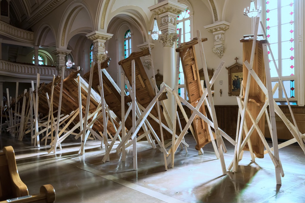Détail d'une installation sculpturale dans une église. Les bancs sont soulevés à l'aide de bois d'œuvre pour former une grande vague.
