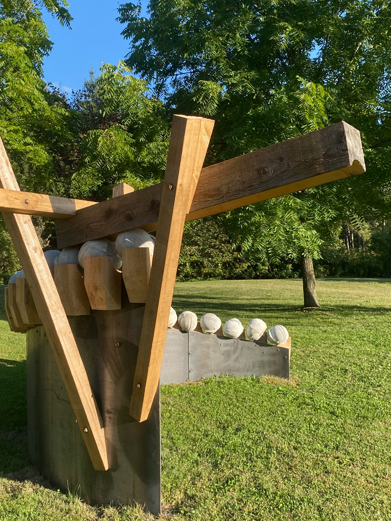 Sur ce qui semble être un roulement à billes fait de casques de chantier moulés en béton, une poutre de bois excavée poursuit son ascension dans un mouvement arqué.
Détail de la poutre se dégageant en porte-à-faux.