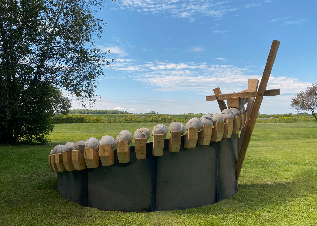 Sur ce qui semble être un roulement à billes fait de casques de chantier moulés en béton, une poutre de bois excavée poursuit son ascension dans un mouvement arqué.