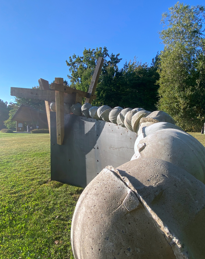 Sur ce qui semble être un roulement à billes fait de casques de chantier moulés en béton, une poutre de bois excavée poursuit son ascension dans un mouvement arqué.
Détail des casques de béton.