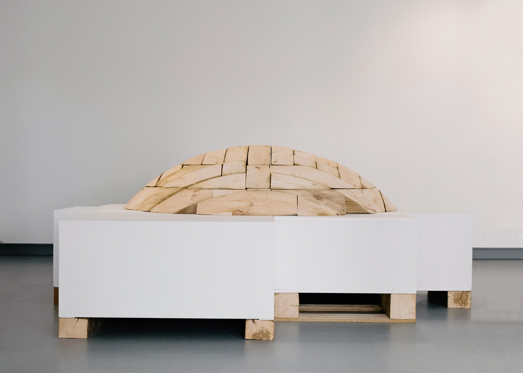Formant un dôme, la sculpture de bois est déposée sur un assemblage de socles dépareillés, lesquels reposent sur des cales.
