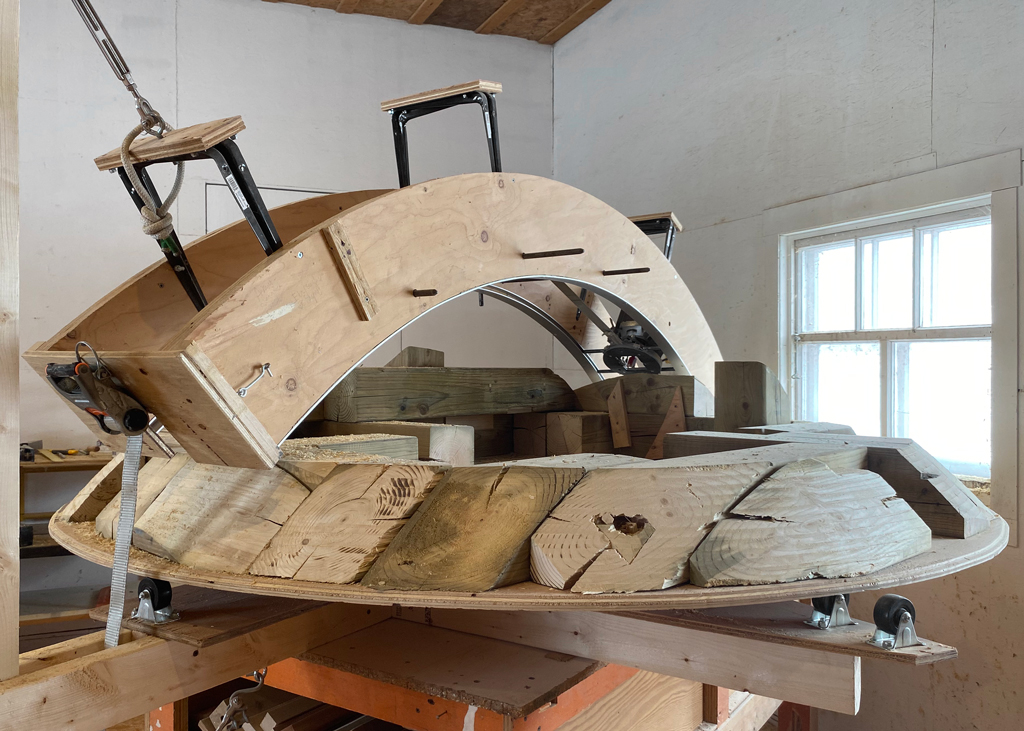 Une grande sculpture de bois en cours de réalisation, enchâssée dans un appareillage servant à l'usiner. Fixée à un plateau tournant, la sculpture est surmontée d'une toupie glissant contre des rails.