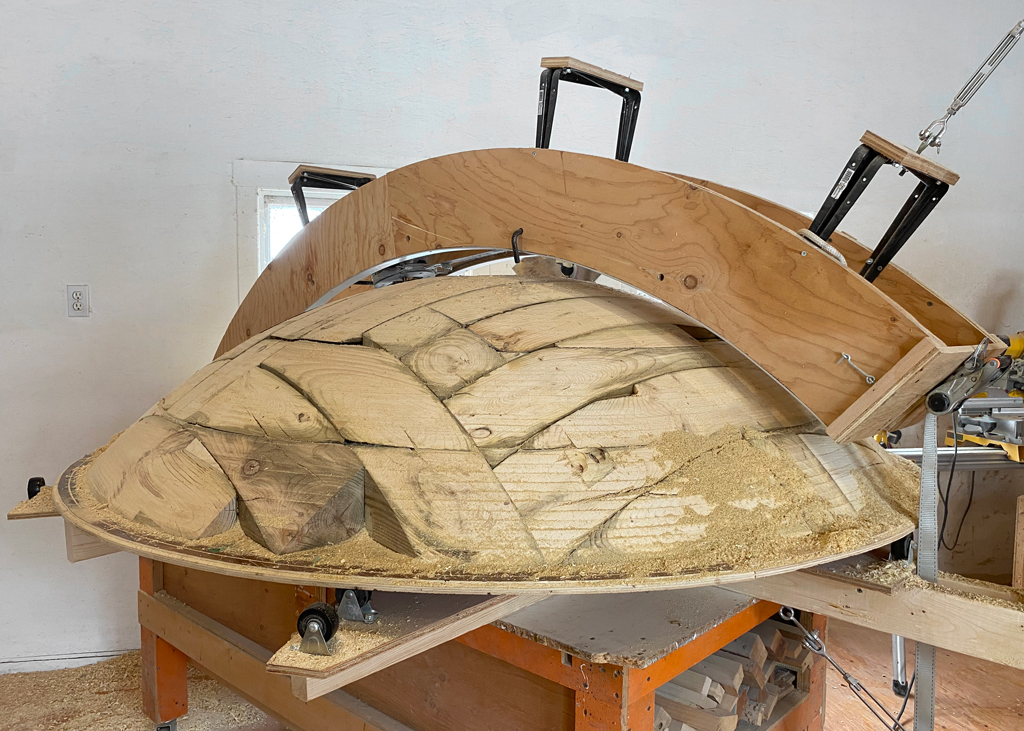 La sculpture de bois, enchâssée dans un appareillage servant à l'usiner vient d'être terminée. Fixée à un plateau tournant, la sculpture est surmontée d'une toupie glissant contre des rails.