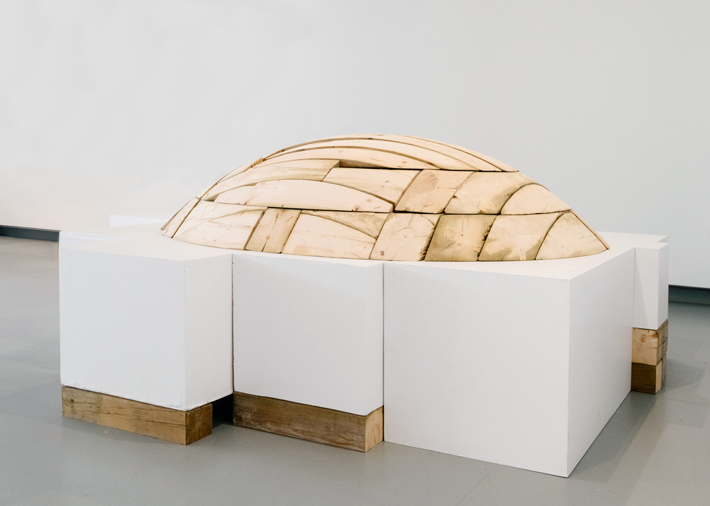 Formant un dôme, la sculpture de bois est déposée sur un assemblage de socles dépareillés, lesquels reposent sur des cales.